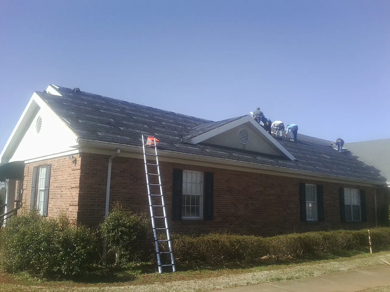 Crew tearing shingles off a brick ranch roof with a ladder set — Metal Roof Repair work by our crew in Cambridge City