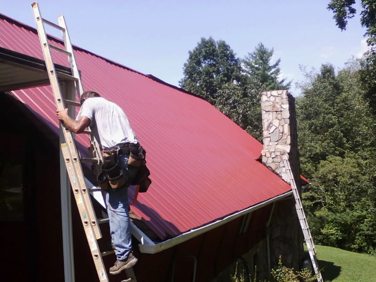 Roofer on a ladder beside a red metal roof and stone chimney — Soffit & Fascia Repair by Roofing Brothers in Cambridge City